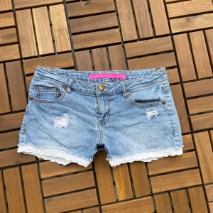 Jeansshorts med spetskant - Snygga ljusblå jeansshorts med slitna detaljer och vit spetskant längst ner. Klassisk femficksmodell med knapp och dragkedja. Perfekta för sommaren! Måtten finns på bilden💗skulle säga att de är storlek s/m💗Kolla gärna in allt annat jag säljer!🥰