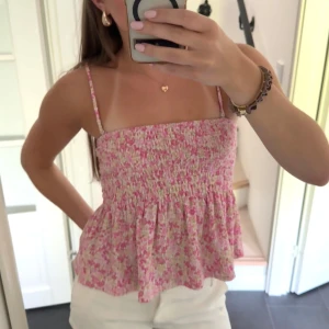 Blommig rosa smockad topp - Supersöt blommig topp i rosa och vita toner med smockad överdel och volangig nederdel. Toppens tunna axelband är avtagbara så du kan bära den som tubtopp också. Perfekt för varma dagar och ger en riktigt somrig vibe.
