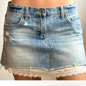 Jeanskjol med spetskant  - Snygg ljusblå jeanskjol med slitna detaljer och råa kanter. Kjolen har en vit broderad spetskant längst ner som ger en söt kontrast till den tuffa denimlooken. Klassisk femficksmodell och bälteshällor. Perfekt för dig som gillar en mix av edgy och cute. Storlek S/M