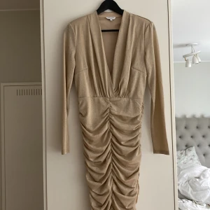 Glittrig champagneklänning Bubbleroom M - Supersnygg glittrig champagnefärgad klänning från Bubbleroom med djup v-ringning, långa ärmar och rynkade detaljer framtill. Klänningen är figurnära och har en kort längd, perfekt för att sticka ut. Materialet är mjukt och stretchigt med metallicskimmer.