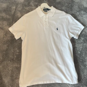 Vit pikétröja från Polo Ralph Lauren - Klassisk vit pikétröja från Polo Ralph Lauren med blå broderad logga på bröstet. Tröjan har korta ärmar, krage och knappar framtill. Tillverkad i bomull och har en snygg custom fit-passform. Perfekt för en clean och stilren look.