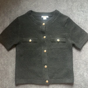 Svart stickad kofta från Gina Tricot - Kortärmad svart stickad kofta från Gina Tricot med snygga guldknappar framtill och två dekorativa fickor på bröstet. Koftan har ribbade kanter vid hals, ärmslut och nederkant. Perfekt för lager-på-lager och en clean look.