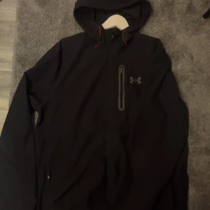 Svart vindjacka från Under Armour L - Svart vindjacka från Under Armour med huva och röda detaljer vid snörningen. Jackan har en bröstficka med dragkedja och Under Armour-logga på bröstet. Perfekt för blåsiga dagar och sportiga outfits.
