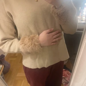 Beige tröja med pälsdetaljer på ärmar - Säljer en beige tröja med rund hals och långa ärmar. Ärmsluten har fluffiga pälsdetaljer i matchande beige ton som ger en unik och mysig look. Perfekt för dig som vill ha något extra till din outfit.