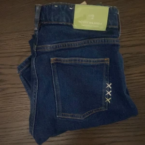 Blå slim jeans från Scotch & Soda - Snygga blå slim jeans från Scotch & Soda med klassisk femficksdesign och coola broderade kryss i vitt, blått och grönt på bakfickan. Jeansen är i stretchig bomull och har en normal passform med smal siluett. Perfekta för en trendig vardagslook. Kom med frågor eller bud! 