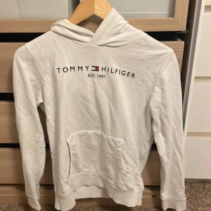 Vit hoodie från Tommy Hilfiger - Vit hoodie från Tommy Hilfiger med klassisk logga och text på bröstet. Tröjan har huva, känguruficka och långa ärmar. Perfekt för en chill och stilren look. Mjuk bomullskänsla och enkel att matcha med jeans eller joggers. Hoodien har en fläck på tröjan men syns inte från distans, väldigt bra skick och perfekt för ert barn som fortfarande växer och inte vill spendera mycket pengar