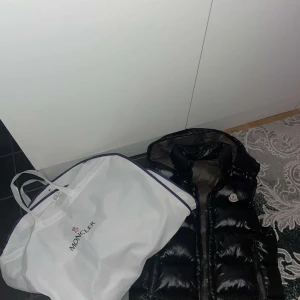 Moncler väst - Säljer en svart, glansig dunväst från Moncler med hög krage och dragkedja framtill. Västen har två fickor med dragkedja och ikonisk Moncler-logga på bröstet. Insidan är grå och quiltad för extra värme. Perfekt för lager-på-lager-stil. Passar även M möts o Sthlm 