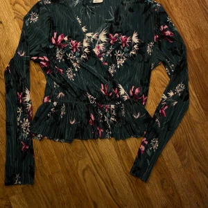 Turkos omlottopp - Supersnygg omlottopp från Gina Tricot i turkos med blommigt mönster i rosa och vit. Toppen har långa ärmar, volangkant nertill och  omlott-skärning. Den är även smått genomskinlig men det löser man lätt med en enkel topp under! Den har en skada i tyget bak, men det är inget hål och är knappt synlig. Materialet är lätt och känns luftigt, perfekt nu inför sommaren. 💙