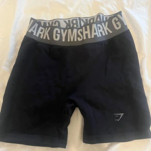 Svarta Gymshark träningstights-shorts - Svarta tighta shorts från Gymshark med bred grå resår i midjan och stor logga. Sitter högt i midjan och är perfekta för gymmet eller löpning. Materialet är stretchigt och andas, vilket gör dem riktigt bekväma under träning. Aldrig använda