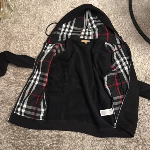 Svart Burberry hoodiejacka M - Svart hoodiejacka från Burberry med klassiskt rutigt foder i huvan och insidan. Jackan har dragkedja, snörning i huvan och två fickor framtill. Ikonisk Burberry-logga på bröstet och snygga detaljer. Perfekt för dig som vill ha en stilren streetlook.HELT NY HELT OANVÄND anledning att jag säljer eftersom att den inte passade funkar för M/S 