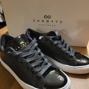 Svarta sneakers från Sandays, strl 37 - Svarta sneakers från Sandays Footwear med glansig finish och vita platta sulor. Skorna har snörning och rund tå, samt detaljer i svart skinnliknande material. Perfekta för en clean och stilren look. Nyskick! Levereras i originalkartong med extra snören.