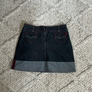 Mörk jeanskjol från MNG Denim - Snygg mörkblå jeanskjol från MNG Denim med kontrastsömmar i rött och coola fickor fram. Kjolen har bälteshällor, dragkedja och en bredare ljusare kant längst ner. Perfekt till sneakers eller boots för en chill look.