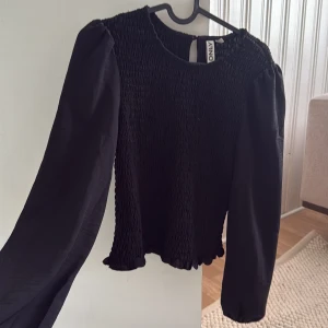 Svart blus med puffärm från ONLY - Supersnygg svart blus från ONLY med smockad kropp och långa puffärmar i tunt tyg. Rund halsringning och liten volangkant nertill. Perfekt för dig som gillar en stilren men ändå trendig look. Använd 1 gång 