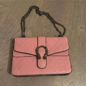 Rosa axelväska med kedja - Säljer en snygg rosa axelväska  med präglat GG-mönster och coolt spänne framtill. Väskan har en robust kedjerem i metall och är gjord i skinn. Perfekt storlek för det viktigaste och ger en lyxig touch till din outfit.