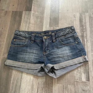 Lågmidjade jeansshorts  - Snygga blå jeansshorts från Jennyfer med uppvikta benslut och flätad detalj i midjan. Klassisk femficksmodell med dragkedja och cool knapp framtill. Perfekta för sommaren och lätta att matcha med allt. Broderad text på insidan av linningen ger en extra touch. Dragkedjan är sönder men går kanske att fixa!!!!! ⚠️ BARA POSTNORD!!