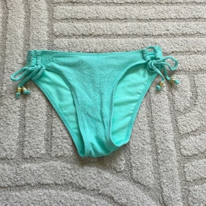 Turkosa bikinitrosor med snörning - Snygga turkosa bikinitrosor med diskret mönster och knyt i sidorna med små gulddetaljer. Perfekta för stranden eller poolen, med en fräsch och ungdomlig vibe.
