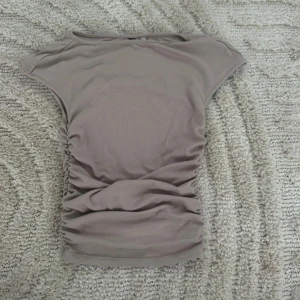 Beige ärmlös topp med rynkade sidor - Snygg beige topp med rynkade detaljer längs sidorna och ärmlös design. Mjuk och stretchig bomullskänsla, perfekt för en stilren och enkel look. Rund halsringning och figurnära passform som framhäver siluetten.