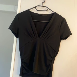 Svart v-ringad topp från BikBok  - Svart topp från BikBok i storlek XS med djup v-ringning och korta ärmar. Snyggt rynkad vid bysten och midjan för en cool siluett. Mjuk och stretchig känsla, perfekt till jeans eller kjol. Enkel men ändå trendig design.
