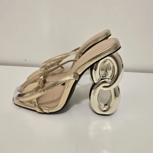 Guldiga sandaler med unik klack - Snygga sandaler i metallic guld med tunna remmar och fyrkantig tå. Klacken är riktigt unik med en chunky design i form av sammankopplade ringar. Perfekta för dig som vill sticka ut och älskar statement-detaljer.