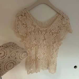 Beige virkad blus med spetsdetaljer - Superfin beige blus i virkad spets med blommigt mönster och korta ärmar. Den är genombruten och har en rund halsringning. Perfekt att styla över ett linne eller bikini för en chill sommarlook.