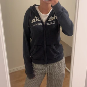 Washed mörkblå hoodie från Hollister - Mysig mörkblå hoodie från Hollister med vit broderad logga på bröstet. Tröjan har huva med snörning, dragkedja framtill och två stora fickor. Perfekt för chill dagar och enkel att matcha med jeans eller mjukisbyxor. Orginalpris är 579kr