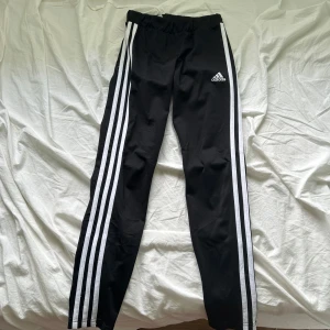 Svarta Adidas byxor med vita ränder - Svarta Adidas byxor med klassiska tre vita ränder längs sidorna och Adidas-logga på låret. Byxorna har resår i midjan och är tillverkade i ett mjukt och stretchigt material, perfekt för träning eller chill. Primegreen-material.