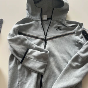 Grå Nike tech set  - Snygga grå mjukisbyxor från Nike med svart logga på benet. Byxorna har dragsko i midjan och muddar vid bensluten för en skön och avslappnad passform. Perfekta för chill eller träning. Tillverkade i mjukt bomullsmaterial. OBS! Hoodien storlek S och byxorna storlek M