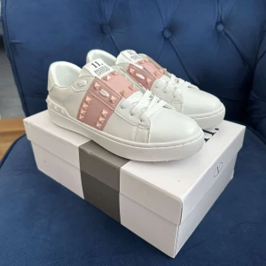 Valentino Rockstud sneakers vit/rosa - Snygga Valentino Garavani Rockstud sneakers i vitt skinn med ljusrosa band och ikoniska nitar. Klassisk rund tå, platt sula och vita snören. Perfekt för dig som vill ha en lyxig och trendig look med extra edge.