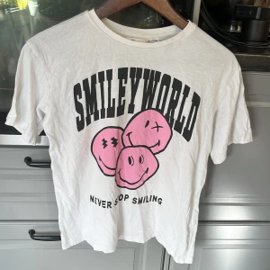 Vit SmileyWorld t-shirt från H&M - Cool vit t-shirt från H&M med stort SmileyWorld-tryck i svart och rosa på framsidan. T-shirten har korta ärmar, rund hals och är gjord i mjuk bomull. Perfekt för dig som gillar statement-plagg och färgglada prints med två fläckar 