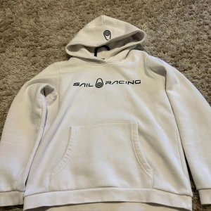 Vit hoodie från Sail Racing - Vit hoodie från Sail Racing med svart logga på bröstet och liten logga på huvan. Klassisk känguruficka framtill och mjuk insida. Perfekt för dig som gillar stilren och sportig look. Långärmad och med huva för extra komfort.