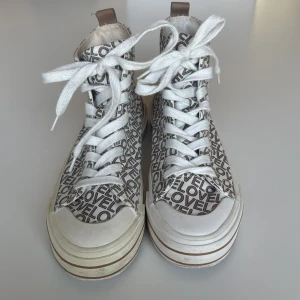 Vita sneakers med LOVE-print från Graceland - Höga vita sneakers från Graceland med beige LOVE-tryck över hela skon. Snygga vita snören, bruna detaljer vid häl och sula samt rund tå. Ovandel i textil och sula i gummi. Perfekta för dig som gillar statement-skor med coolt mönster.