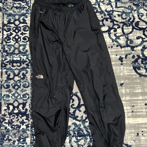 Svarta täckbyxor från The North Face - Svarta täckbyxor från The North Face med HyVent-teknologi. Byxorna har elastisk midja, meshfoder och dragkedjor vid fickorna. Tillverkade i nylon med polyesterfoder, perfekta för kalla och blöta dagar. Snygg logga broderad på benet.