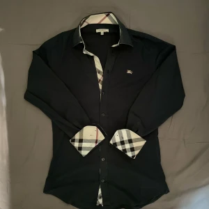 Burberry skjorta storlek M  - Svart skjorta från Burberry med klassiska rutiga detaljer på insidan av kragen och manschetterna. Skjortan har lång ärm, knappar framtill och liten broderad logga på bröstet. Tillverkad i bomull med lite stretch för extra komfort. Mycket bra skick men är lite sliten i kragen (se bild 5)⭐️⭐️