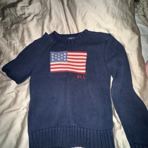 Mörkblå stickad tröja Polo Ralph Lauren - Snygg mörkblå stickad tröja från Polo Ralph Lauren med amerikansk flagga framtill och RL-broderi. Klassisk rund halsringning och långa ärmar. 