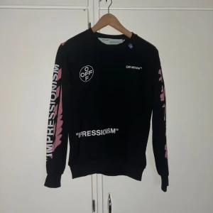 Off-White svart sweatshirt med tryck - Svart sweatshirt från Off-White med vita och rosa tryckdetaljer. Framsidan har logga och text, medan baksidan har det ikoniska Off-White-korset i rosa. Långa ärmar med text och rosa grafiska detaljer. Crewneck och ribbade muddar. Perfekt för dig som gillar streetwear.