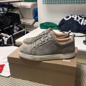 Grå mockasneakers från Louboutin - Snygga grå sneakers i mocka från Christian Louboutin med klassisk röd sula och vita kanter. Skorna har rund tå, snörning och diskreta detaljer. Insidan är ljusbeige och de har en stilren, minimalistisk look som passar till det mesta.