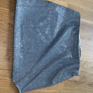Glittrig silverkjol från Noisy May - Supersnygg kort glittrig kjol från Noisy May i silver. Kjolen har ett diskret rutigt mönster och dragkedja bak för enkel påklädning. Perfekt för dig som vill sticka ut med lite extra bling. Sitter tajt och har en modern vibe.