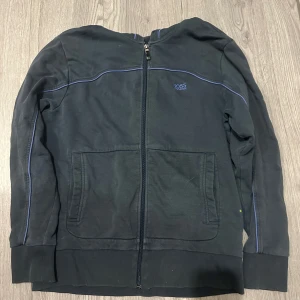 Svart zipjacka från Hugo Boss - Mörk blå zip kofta från Hugo Boss med blå detaljer och broderad logga på bröstet. Koftam har långa ärmar, ribbade muddar och två stora fickor framtill. Tillverkad i mjuk bomull, perfekt för en chill och stilren look.