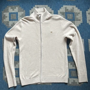 Beige kofta från Jack & Jones - Säljer en stilren beige kofta från Jack & Jones med hel dragkedja och hög krage. Tröjan är stickad i bomull och har ett diskret broderat emblem på bröstet. Perfekt för lager-på-lager och passar till många olika outfits. Tröjan är även helt oanvänd.  