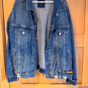 Zara jeansjacka med tryck bak - Snygg blå jeansjacka från Zara med coolt grafiskt tryck och text på ryggen. Jackan har klassiska bröstfickor med knappar, slitna detaljer nertill och en relaxed fit. Perfekt för dig som vill sticka ut med streetstyle-vibe.