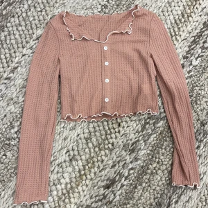 Rosa croppad blus med volangkant - Supersöt rosa croppad blus med långa ärmar, vita knappar framtill och volangkant längs krage, ärmslut och nederkant. Storlek S