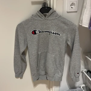 Grå Champion hoodie med logga - Säljer en grå hoodie från Champion med stor broderad logga över bröstet och liten logga på ärmen. Klassisk känguruficka framtill, huva och ribbade muddar. Perfekt för chill dagar eller när du vill ha en enkel streetstyle-look. Endast använd 3 gånger.