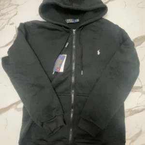Svart hoodie från Polo Ralph Lauren - Svart hoodie från Polo Ralph Lauren med dragkedja, huva och snörning. Klassisk vit broderad logga på bröstet och ribbade muddar. Perfekt för chill dagar och enkel att matcha med jeans eller joggers.
