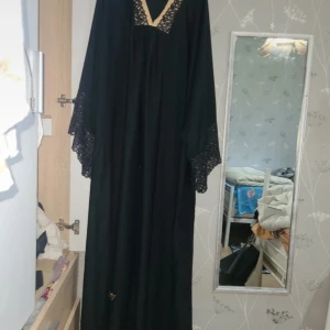 Svart långklänning med spetsdetaljer - Svart långklänning med vida ärmar och snygga spetsdetaljer vid ärmslut och v-ringad hals. Guldigt broderi nertill och vid halsen ger en lyxig touch. Klänningen har en loose passform och faller snyggt. Perfekt för dig som gillar elegant och stilren look.