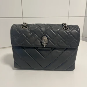 Kurt Geiger väska - Snygg grå handväska från Kurt Geiger i quiltat skinn med silverfärgad kedjerem i den större modellen. Uppriven på tyget inuti och därav priset