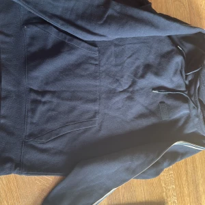 Svart hoodie med huva och ficka - Svart hoodie med klassisk känguruficka framtill och dragsko i huvan. Tröjan har långa ärmar och en enkel, stilren design utan tryck. Perfekt för chill dagar eller när du vill ha något bekvämt och snyggt.