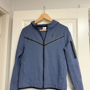 Blå Nike Teck Hoodie - Nike Tech hoodiejacka i blåbärsfärg med hel dragkedja och luva. Snygg V-detalj framtill, svarta detaljer och ficka med dragkedja på ärmen. Perfekt för chill dagar eller när du vill ha en sportig look.