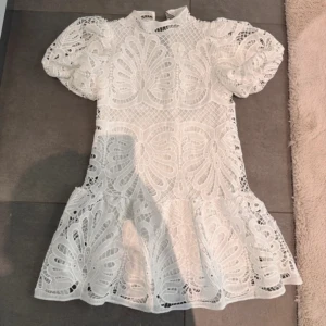 Vit spetsklänning med puffärm - Superfin vit kortklänning i spets med unikt broderat mönster och volang nertill. Klänningen har puffiga korta ärmar och hög hals. 