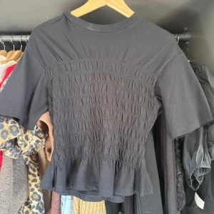 Svart smockad topp med volangkant - Supersnygg svart topp från GINA med smockad midja och volangkant nedtill. Modellen har korta ärmar och en tightare passform över kroppen. Perfekt att styla med jeans eller kjol för en trendig look.