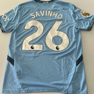 Manchester City Savinho matchtröja Puma - Manchester Citys matchtröja med Savinho 26-tryck. Ljusblå med vita detaljer, Premier League-märke i guld på ärmen och klubbmärke framtill. Tillverkad i ventilerande polyester med korta ärmar och snygga svarta inslag på sidorna. Endast handtvätt gäller. Replika